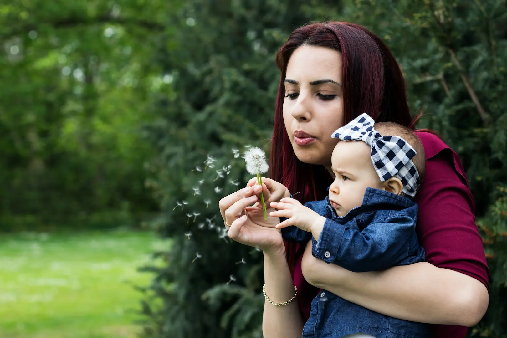 A woman holding a baby and blowing on a dandelion.