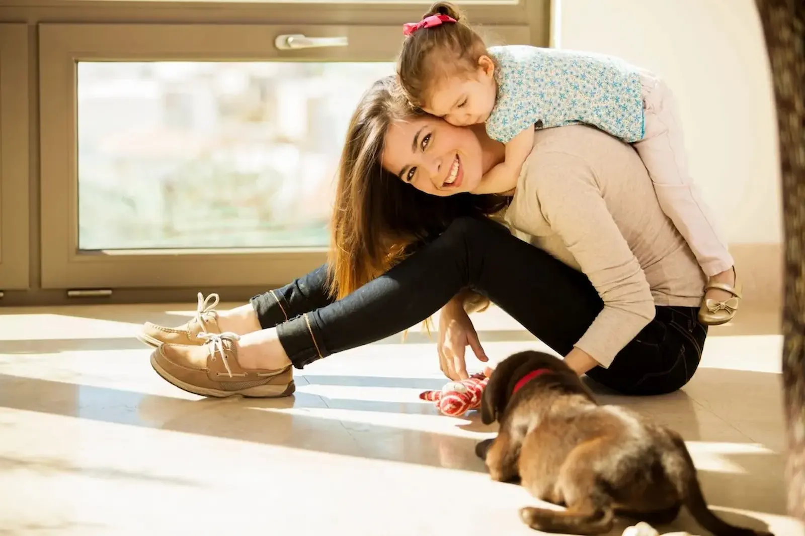 nanny child sitting on the floor with her young daughter on her back and a puppy playing beside them